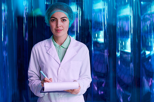 Waist up portrait of smiling female doctor  and holding clipboard while posing against blue plastic wall, copy space