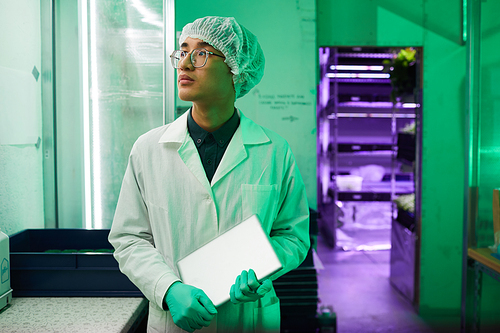 Waist up portrait of young Asian worker wearing protective clothing standing in bio laboratory lit by green light, copy space