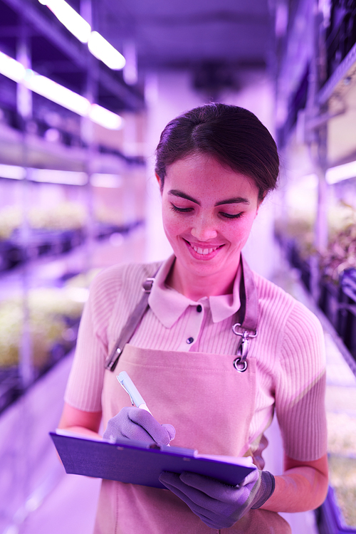 Waist up portrait of smiling female worker writing on clipboard while standing in  greenhouse lit by UV light, copy space