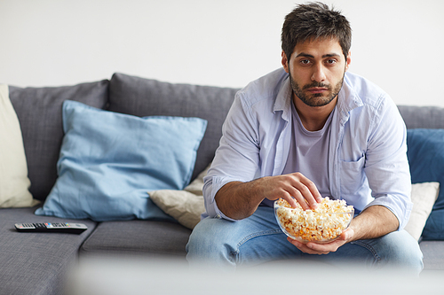Portrait of sad bearded man watching TV and holding bowl of popcorn while sitting on sofa at home, copy space