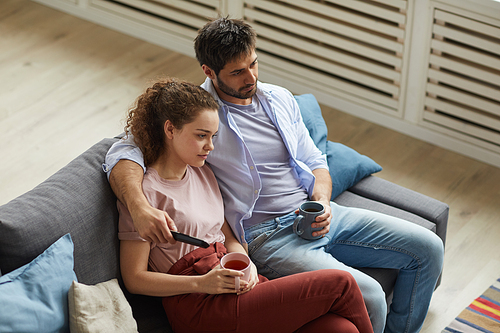 High angle portrait of modern couple watching TV and holding mugs while sitting on sofa at home in cozy apartment and enjoying  time, copy space