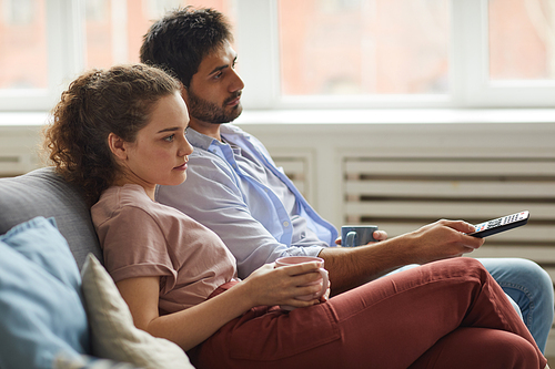 Side view portrait of modern couple watching TV and holding mugs while sitting on sofa at home in cozy apartment, copy space