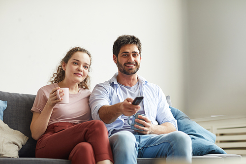Portrait of modern couple watching TV and holding mugs while sitting on sofa at home in cozy apartment enjoying  time, copy space