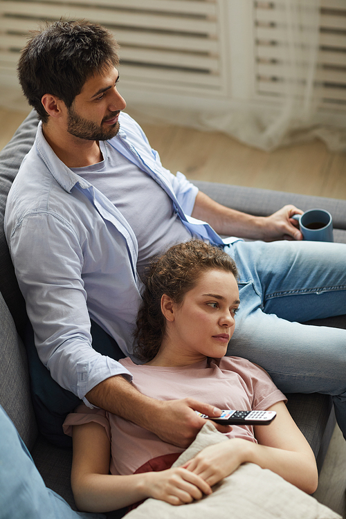 High angle view at modern couple watching TV together while lying on sofa at home in cozy apartment enjoying  time, copy space