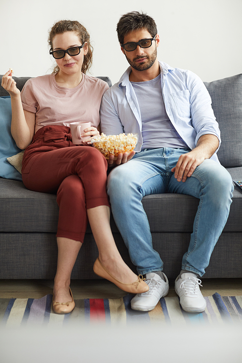 Vertical full length portrait of modern couple watching TV together while sitting on sofa and wearing sunglasses at home enjoying  time
