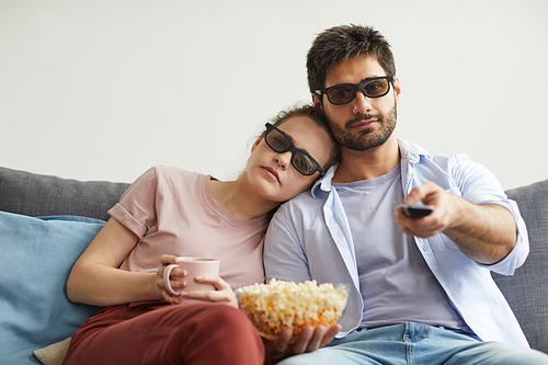 Portrait of modern couple watching TV together while sitting on sofa and wearing stereo glasses at home enjoying  time, copy space