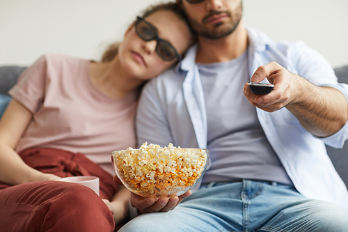 Blurred portrait of modern couple watching TV with stereo glasses while holding bowl of popcorn, focus on foreground, copy space