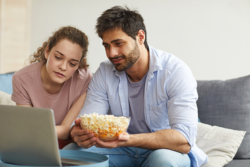 Portrait of modern young couple watching movies online via laptop and holding big bowl of pop corn, copy space