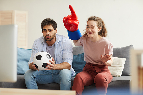 Couple of enthusiastic sport fans watching football match on TV at home and cheering emotionally, focus on young woman wearing foam finger, copy space