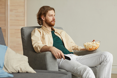 Portrait of modern bearded man watching TV at home while relaxing in comfy armchair and holding bowl of popcorn, copy space