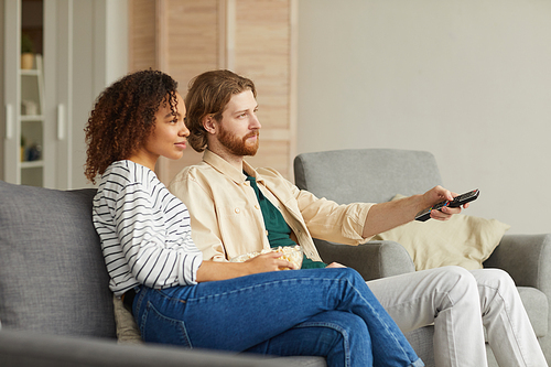 Side view portrait of modern mixed-race couple watching TV at home while relaxing on cozy sofa, focus on beaded man holding remote control, copy space