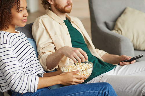 Cropped portrait of modern mixed-race couple watching TV at home while relaxing on cozy sofa, focus on hands holding bowl of popcorn, copy space
