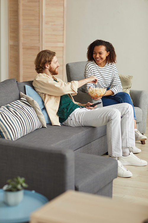 Vertical full length portrait of happy mixed-race couple enjoying time at home, watching TV while relaxing on cozy sofa and eating popcorn, copy space