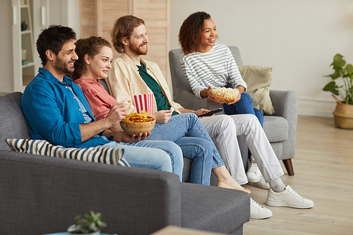 Full length side view at multi-ethnic group of friends watching TV together while sitting on cozy sofa at home and enjoying snacks, copy space