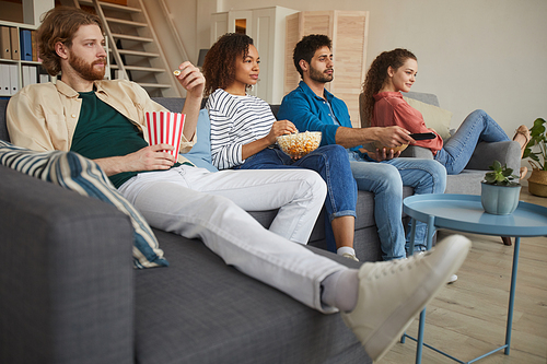 Low angle view at multi-ethnic group of friends watching TV together while sitting on comfortable sofa at home and enjoying snacks, copy space