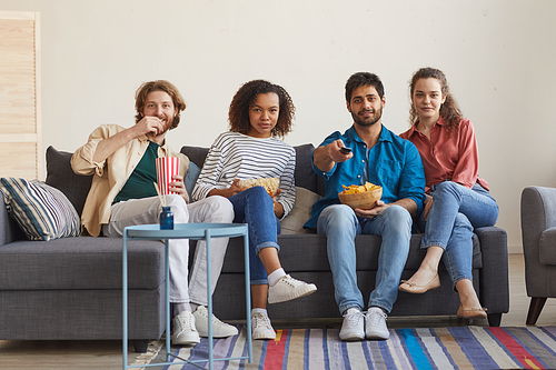 Full length portrait of multi-ethnic group of friends watching TV together while sitting on cozy sofa at home and enjoying snacks, copy space