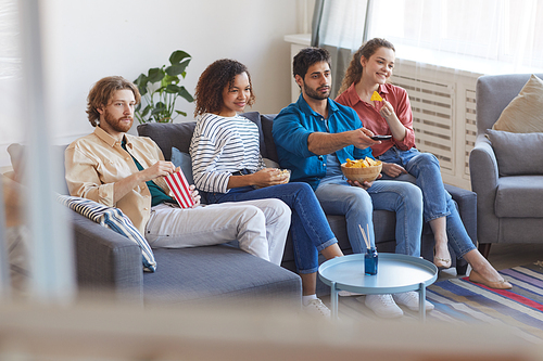 Portrait of multi-ethnic group of friends watching TV together while sitting on comfortable sofa at home and eating snacks, copy space