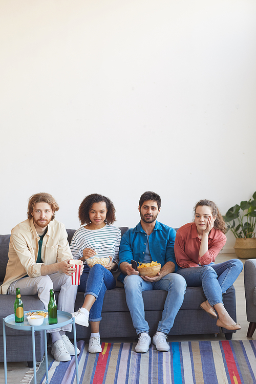 Vertical full length portrait of multi-ethnic group of friends watching TV together while sitting on comfortable sofa at home and enjoying snacks, copy space in top section