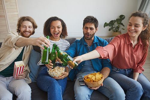Portrait of multi-ethnic group of friends watching TV together while sitting on comfortable sofa at home and clinking beer bottles