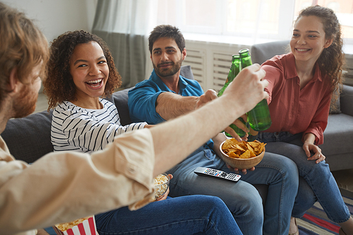 Portrait of multi-ethnic group of friends clinking beer bottles while watching TV together sitting on comfortable sofa at home