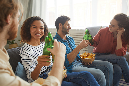 Multi-ethnic group of friends clinking beer bottles while watching TV together sitting on comfortable sofa at home, focus on smiling African-American woman in foreground