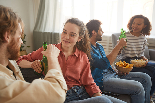 Multi-ethnic group of friends clinking beer bottles while watching TV together sitting on comfortable sofa at home, focus on young couple in foreground