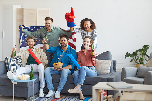 Full length portrait of multi-ethnic group of friends watching sports match on TV and cheering emotionally while holding American flag, copy space