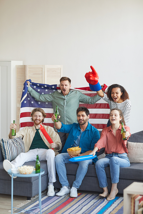 Vertical full length portrait of multi-ethnic group of friends watching sports match on TV and cheering emotionally while holding American flag, copy space