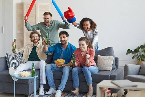 Full length portrait of multi-ethnic group of friends watching sports match on TV and cheering emotionally sitting together on sofa, copy space