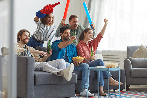 Full length portrait of multi-ethnic group of friends watching sports match on TV and cheering emotionally while sitting together on sofa, copy space