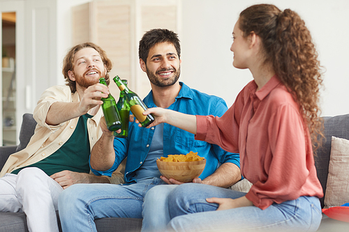 Group of friends clinking beer bottles while watching TV together sitting on comfortable sofa at home, copy space