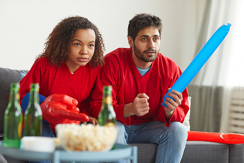 Portrait of modern mixed-race couple watching sports on TV at home and cheering for intense match while wearing red team uniforms, copy space