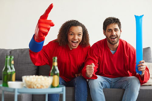 Portrait of modern mixed-race couple watching sports match on TV at home and cheering emotionally while wearing red team uniforms, copy space