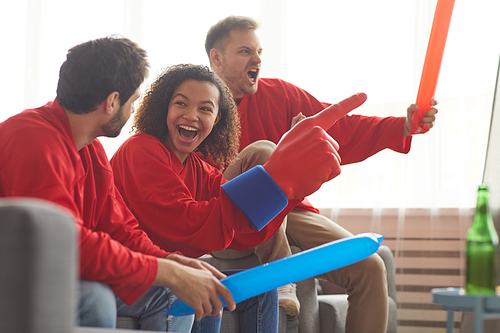 Side view at group of friends watching sports match on TV at home and cheering emotionally while wearing red team uniforms, copy space