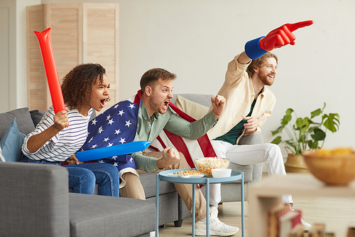 Side view at group of young people watching sports match on TV at home and cheering emotionally while wearing American flag, copy space