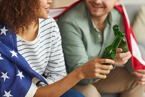 Close up of mixed-race young couple clinking beer bottles while wearing American flag, copy space