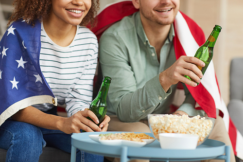 Cropped portrait of mixed-race young couple watching TV at home and drinking beer while wearing American flag, copy space
