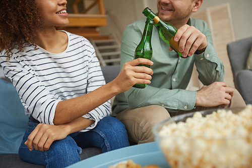 Cropped portrait of modern mixed-race couple watching TV at home and clinking beer bottles while sitting on sofa in cozy apartment