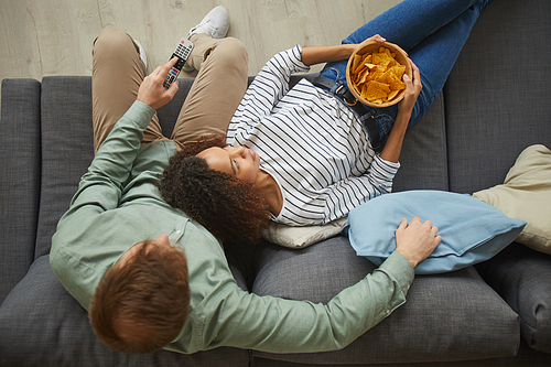 Above view portrait of young mixed-race couple watching TV at home and eating snacks while lying on comfortable sofa, copy space