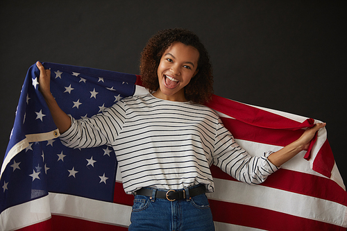 Waist up portrait of young African-American girl holding American flag while posing against black background and  , copy space