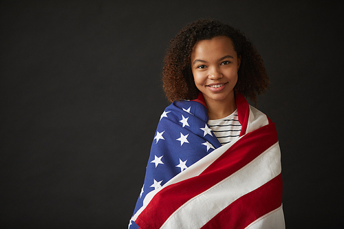 Waist up portrait of young African-American girl wrapped in American flag while standing against black background and smiling at camera , copy space