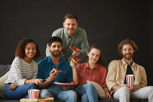 Large group of friends eating pizza and snacks while enjoying party at home sitting on big sofa against black background and all , copy space above