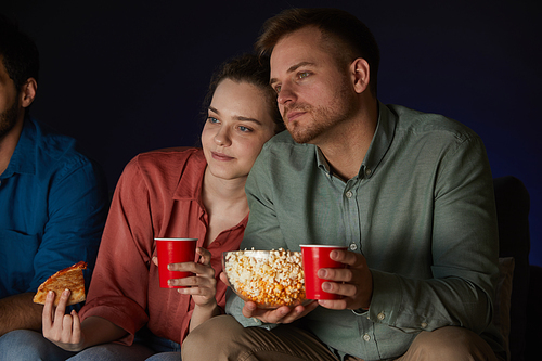 Portrait of adult couple watching movies at home while eating snacks and popcorn sitting on sofa in dark room