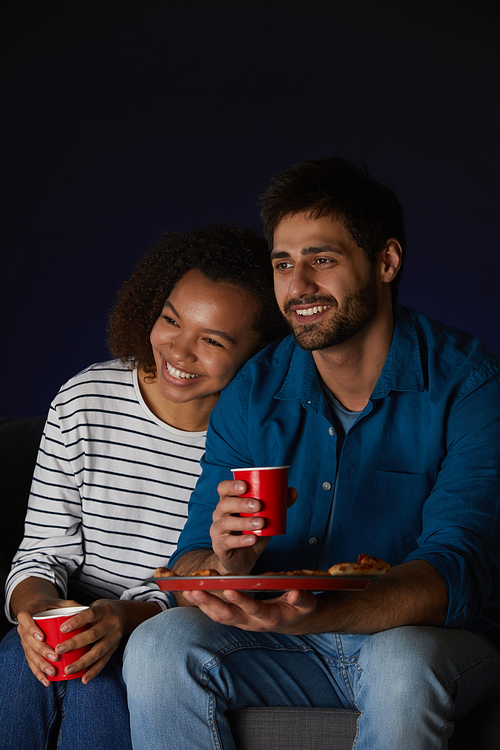 Vertical portrait of mixed-race young couple watching movies at home while eating snacks and popcorn sitting on sofa in dark room