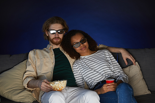 Portrait of mixed-race modern couple watching movie and wearing stereo glasses while sitting on couch in dark room