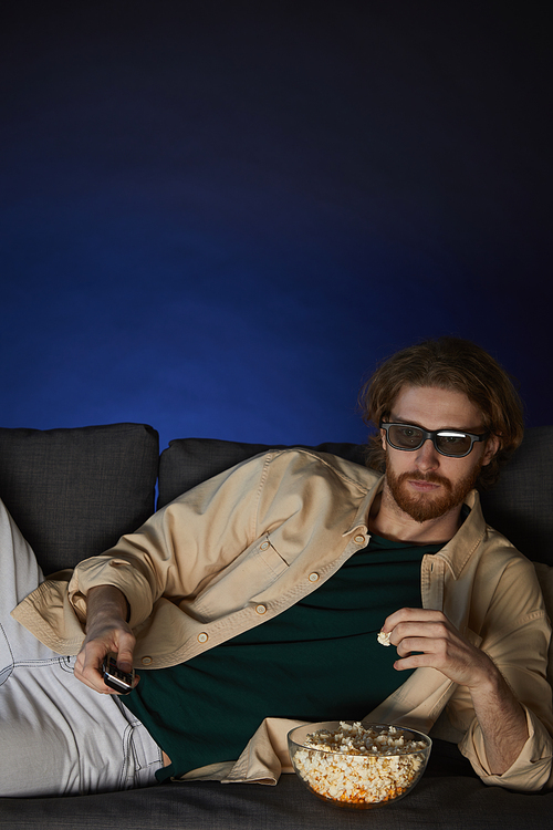 Vertical portrait of modern bearded man watching movie and wearing stereo glasses while lying on couch in dark room, copy space