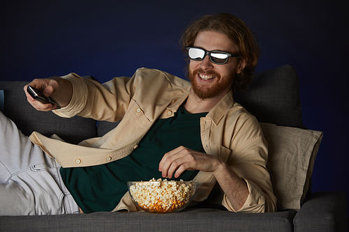 Portrait of modern bearded man watching movie and wearing stereo glasses while lying on couch in dark room, copy space