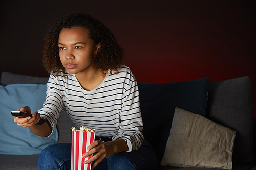 Portrait of young African American woman watching TV at home and holding remote control while sitting on couch in dark room