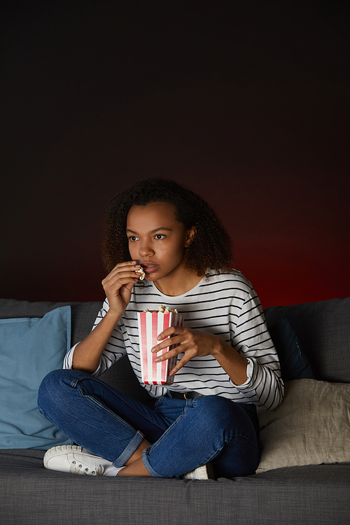 Vertical full length portrait of young African American woman watching TV at home and eating popcorn while sitting cross legged on couch in dark room, copy space