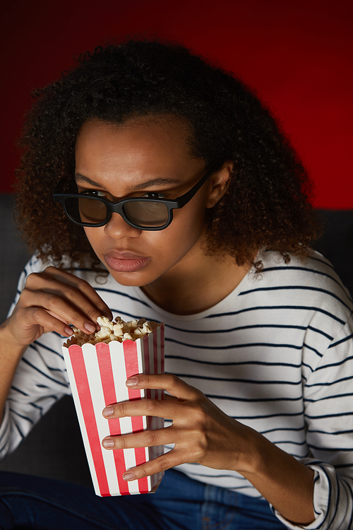 Vertical portrait of young African American woman watching TV at home and eating popcorn while sitting in dark room